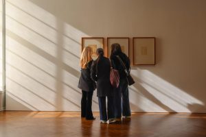 Tres estudiantes observan cuadros en una pared soleada de una galería.