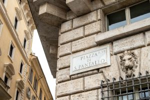 Placa de mármol de la calle Piazza di S. Pantaleo junto a una cabeza de león esculpida en un edificio histórico.
