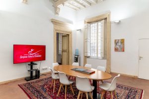 Sala de reuniones con mesa ovalada, sillas blancas, techo de casetones y pantalla con el logo de la European School of Economics.