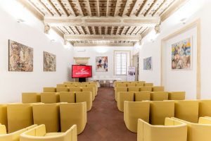 University classroom with rows of yellow armchairs, a coffered ceiling, and a front screen with the European School of Economics logo.