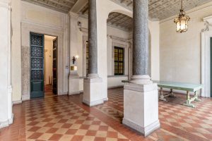 Loggiato storico con grandi colonne in pietra, pavimento a scacchi in cotto, soffitto decorato e un lungo tavolo.