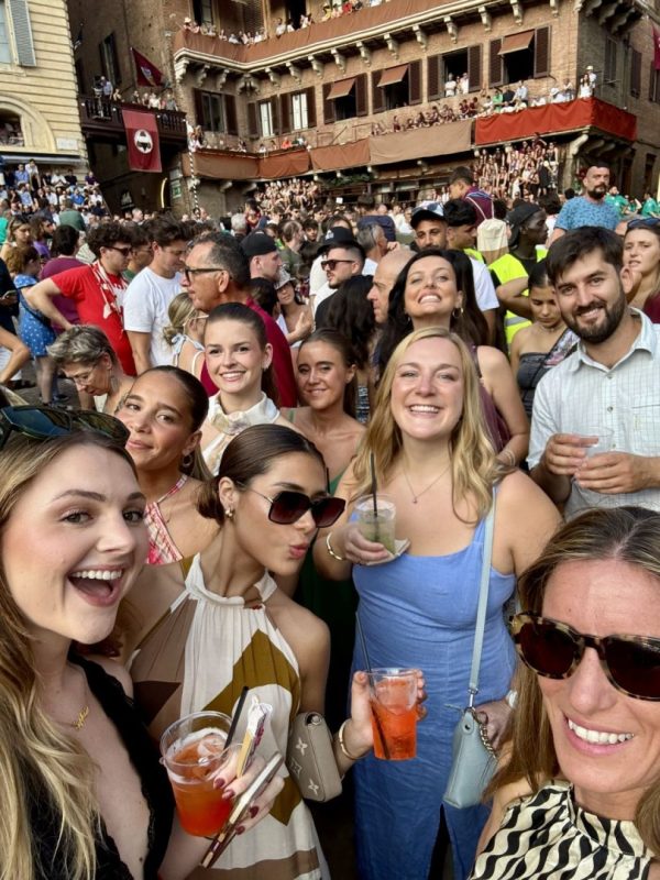 A group of smiling ESE Florence international students taking a group selfie with drinks in hand in Piazza del Campo, surrounded by a lively crowd and historic buildings during the Palio di Siena celebrations.