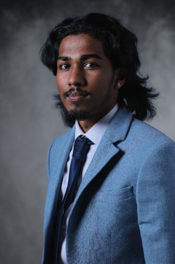 Portrait of Rudi wearing a light blue suit jacket and dark tie against a grey background.